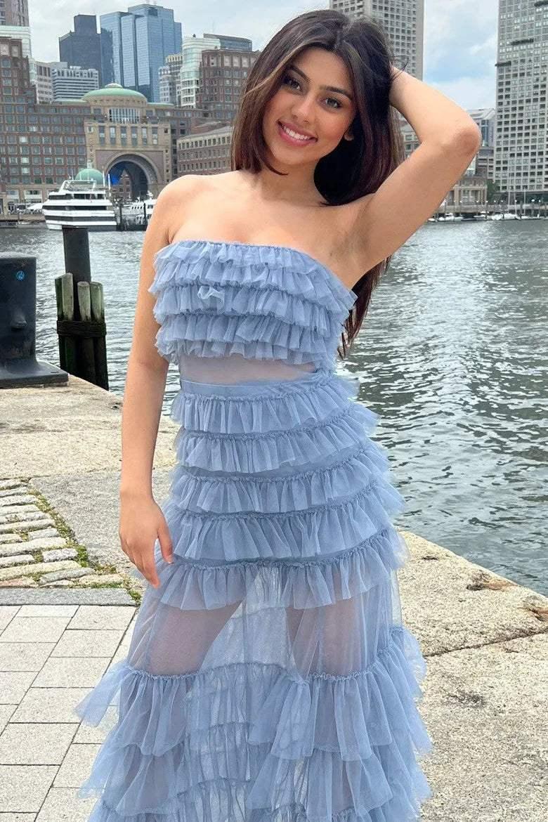 A woman in a blue, ruffled dress stands on a stone walkway near a waterfront. She smiles and poses playfully, with modern buildings and boats in the background.