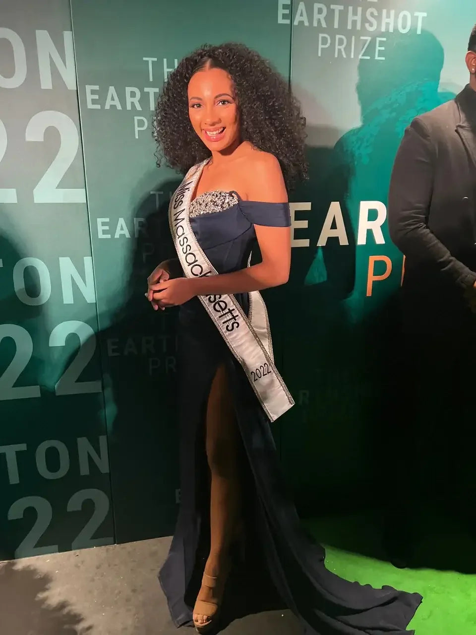 A woman in formal attire poses confidently, smiling. She wears a strapless gown with a high slit and holds a sash that reads "Miss Massachusetts 2022." The background features branding for the Earthshot Prize.