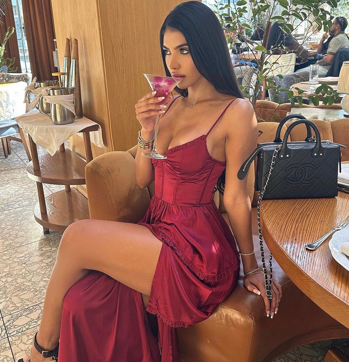 A woman in a red dress sits at a restaurant table, sipping a cocktail. She has long, dark hair and wears jewelry. A black handbag rests nearby on the table.