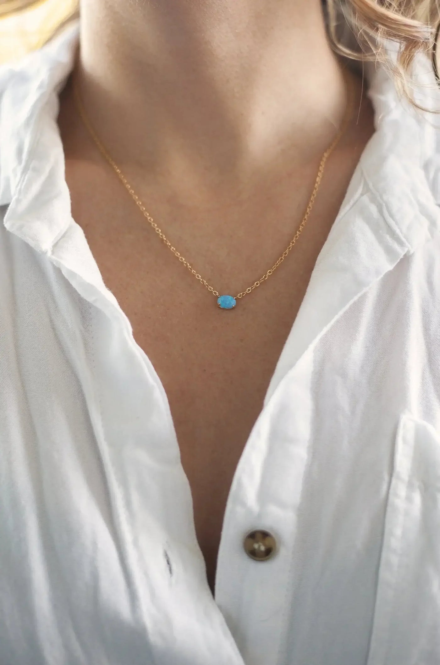 A delicate gold necklace with a blue gem rests on a person's neck, framed by a white shirt. The skin is visible and softly lit, suggesting a casual, relaxed setting.