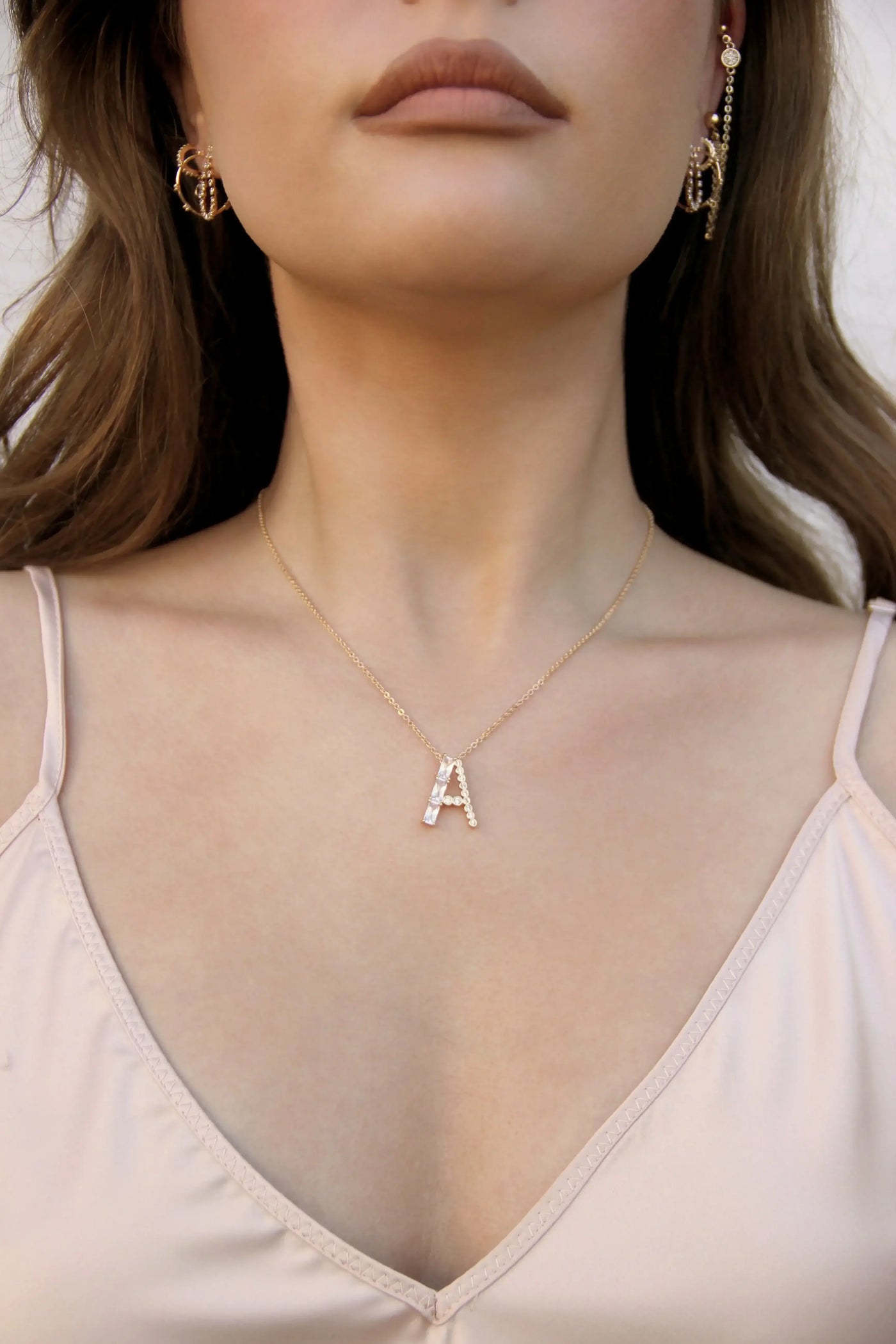 A person wears a gold necklace featuring a decorative letter "A" at the center, paired with dangling earrings. The background suggests an outdoor setting with neutral tones.