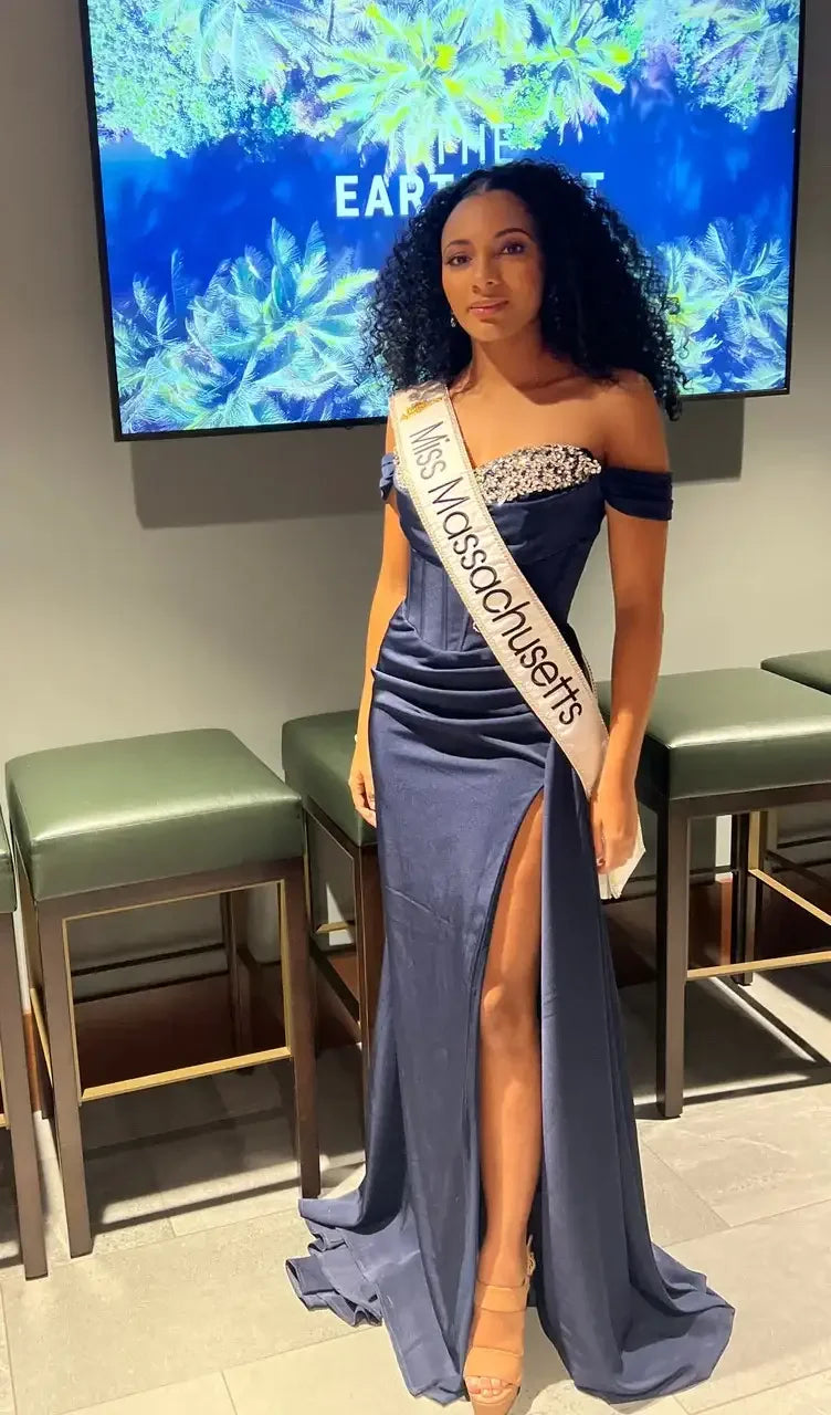 A woman in a navy evening gown stands confidently, displaying a leg slit. She wears a sash that reads "Miss Massachusetts" against a backdrop of tropical foliage and a digital screen.