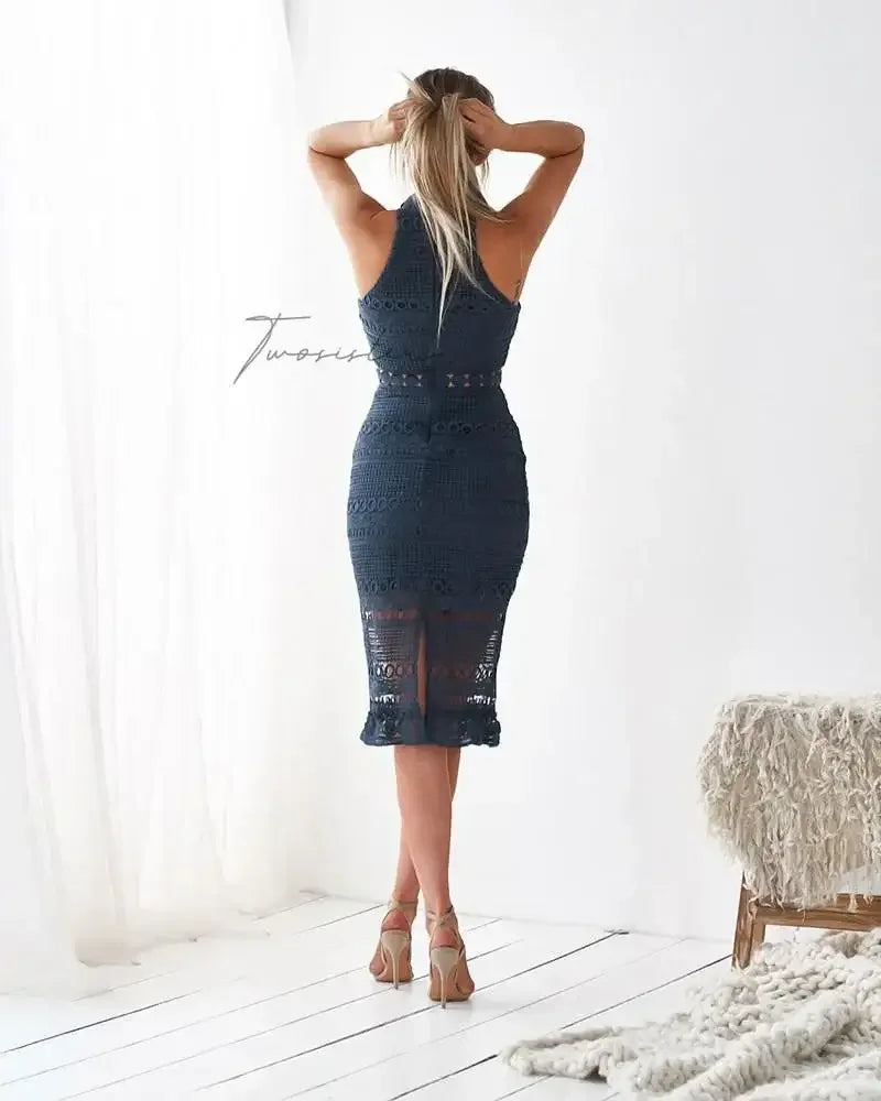 A woman with long hair tied back is pulling her hair up while wearing a fitted dark dress. She stands on white wooden flooring, next to a textured, light-colored chair.