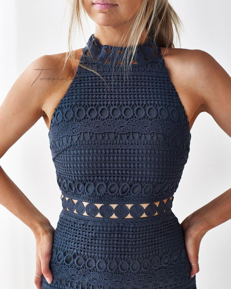 A stylish woman poses confidently with her hands on her hips, wearing a dark sleeveless top featuring intricate patterns. The background is a light, neutral color, enhancing the outfit's details.