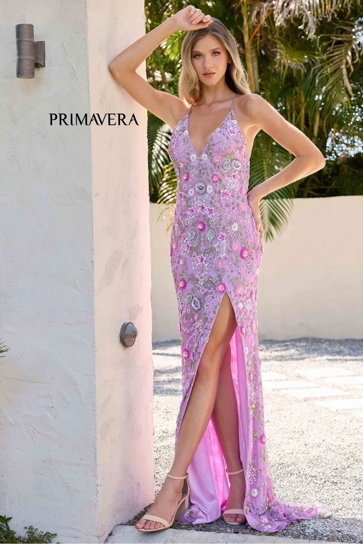 A model wears a lavender pink evening gown with floral embroidery and a high thigh slit, posing outdoors next to a white stucco wall. The brand name 'PRIMAVERA' is visible on the left.