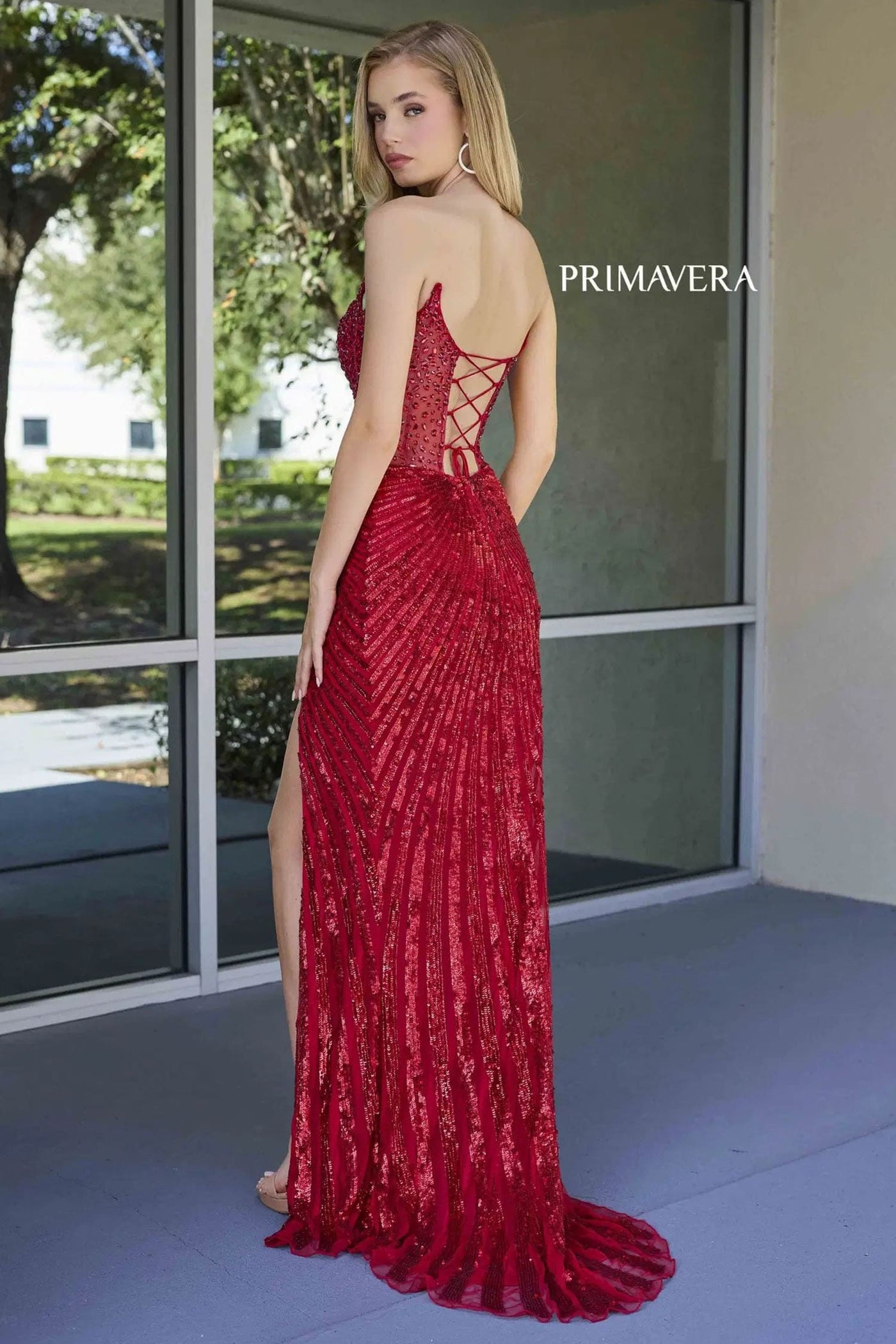 Woman wearing a red lace evening gown with 'Primavera' branding, standing indoors.