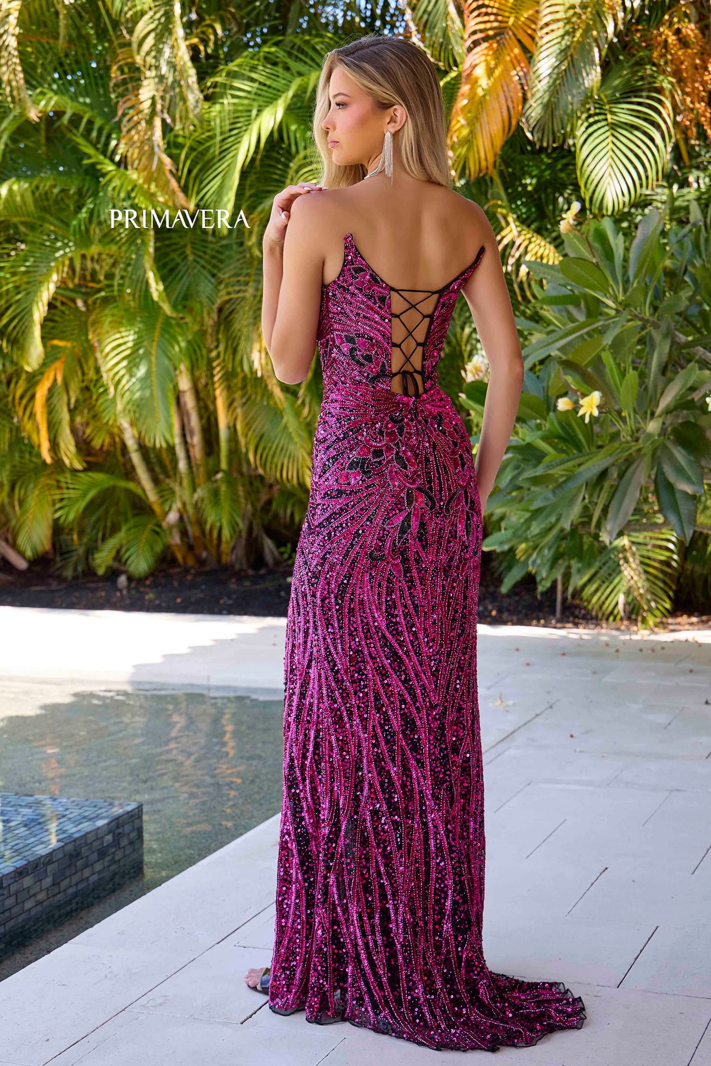 A woman stands outdoors by a pool wearing a long magenta sequined evening gown with thin straps and an open lace-up back. Palm foliage and a patio are visible in the background, and the brand name Primavera appears on the left side.