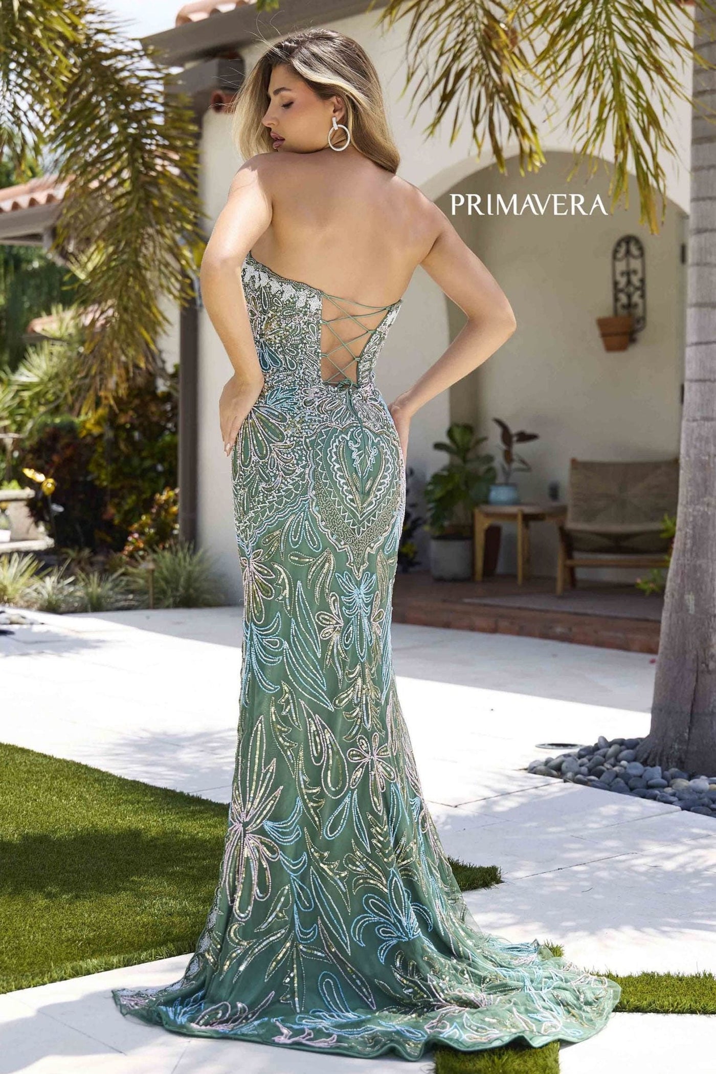 Woman in a green evening gown with a backless design, standing outdoors with palm trees and a house in the background.