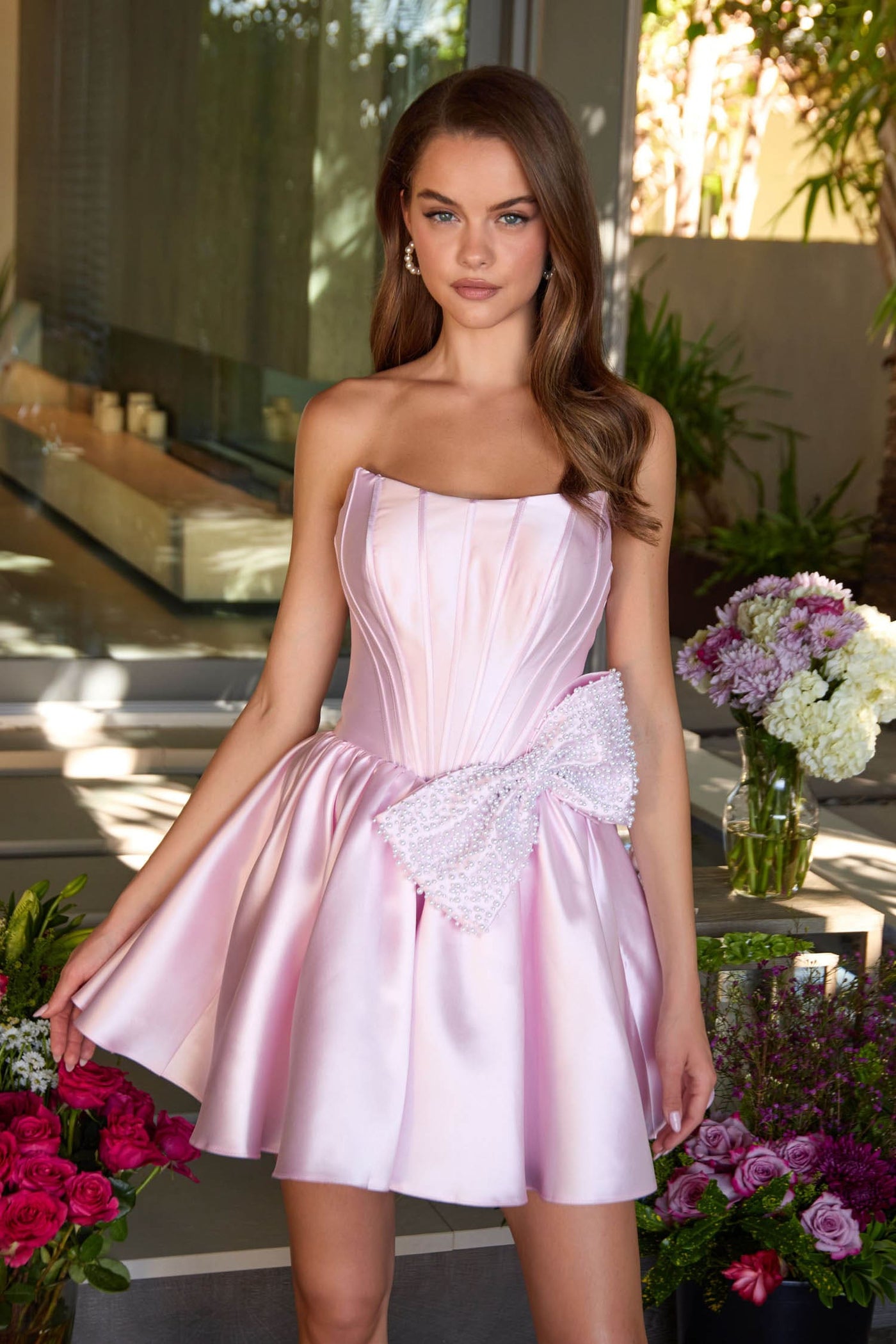 Woman in a pink strapless dress with a bow standing outdoors with flowers around.