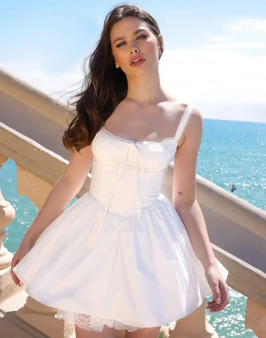 A woman in a white sundress stands by a staircase, gazing directly at the viewer. The background features a sparkling blue ocean under a bright sky, suggesting a sunny day.