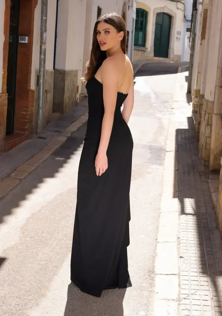 A woman in a strapless black dress stands in a sunlit narrow street, looking over her shoulder with a confident expression, surrounded by old stone buildings.