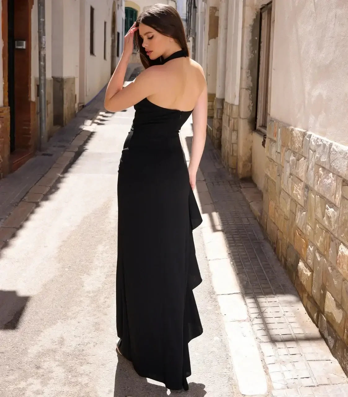 A woman in a black, form-fitting evening gown stands in an alleyway, lightly touching her hair with one hand while looking away, surrounded by white-washed buildings and cobblestone.
