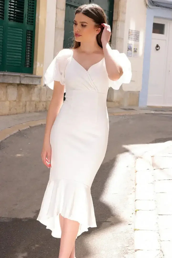 A woman in a white dress with puffed sleeves walks gracefully along a sunlit, narrow street, surrounded by stone buildings and green shutters, showcasing elegance in an outdoor setting.