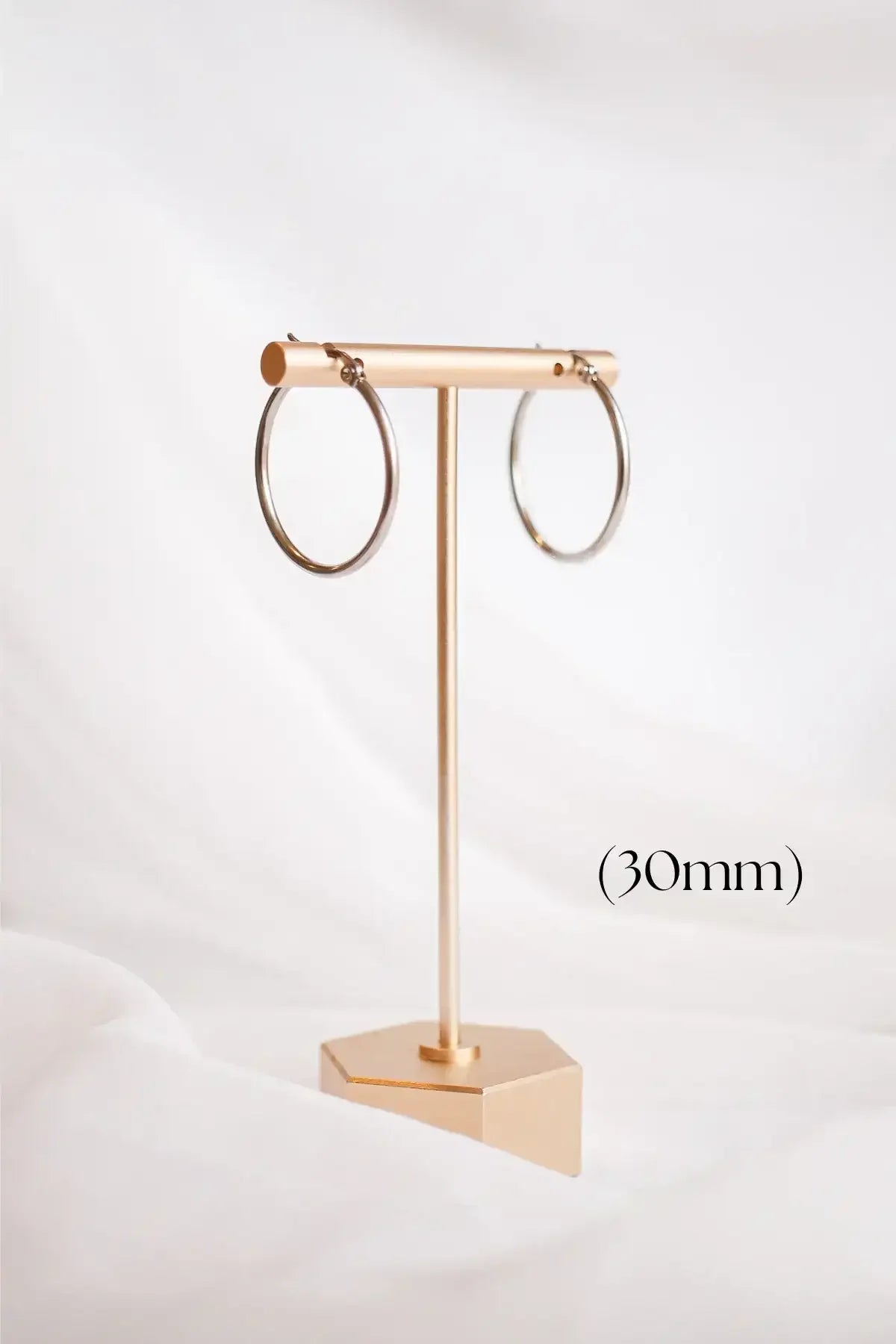 A pair of circular silver hoop earrings hangs on a gold display stand, set against a softly draped white background. (30mm) is noted below.
