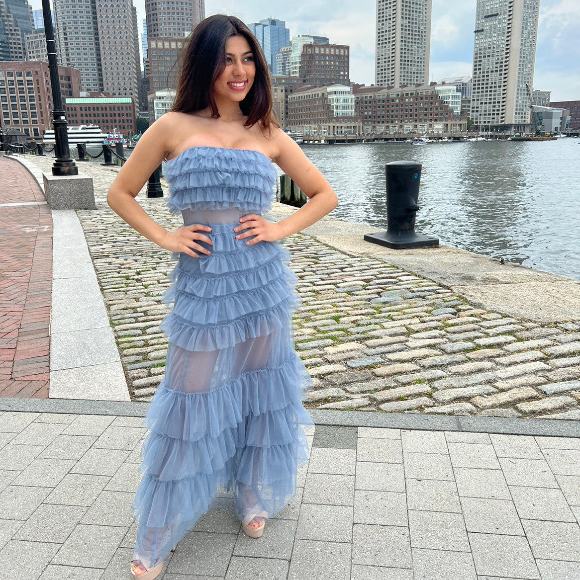 A woman stands by a waterfront in a layered, strapless light blue dress. She's smiling with her hands on her hips, surrounded by cobblestone paths and tall buildings in the background.