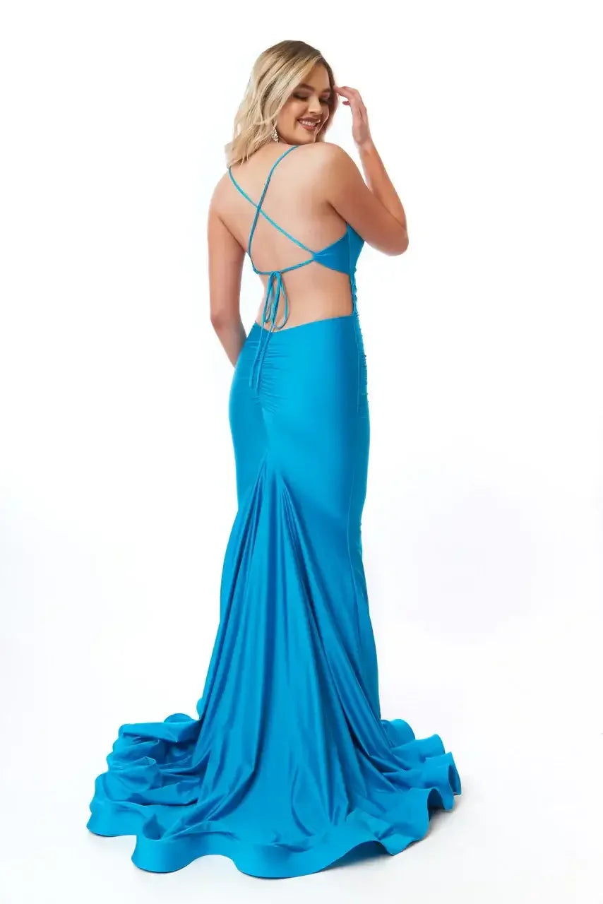 A woman in a vibrant blue gown stands gracefully, showing off the dress's fitted silhouette and dramatic train while playfully touching her hair, against a plain white background.