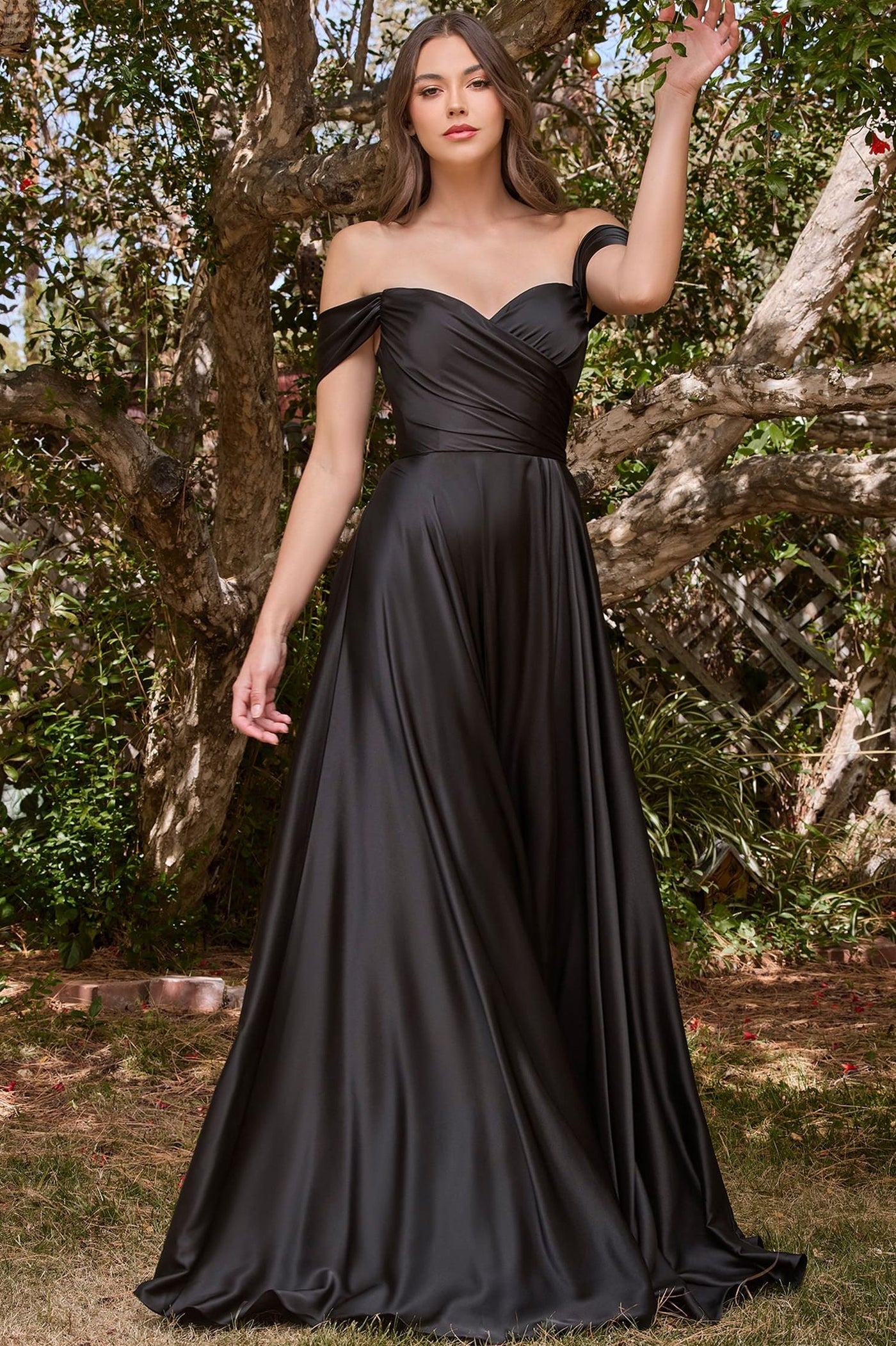 A woman wearing a floor-length off-the-shoulder black satin gown stands outdoors in front of a tree, with one arm raised. The dress has a fitted ruched bodice and a flowing skirt.