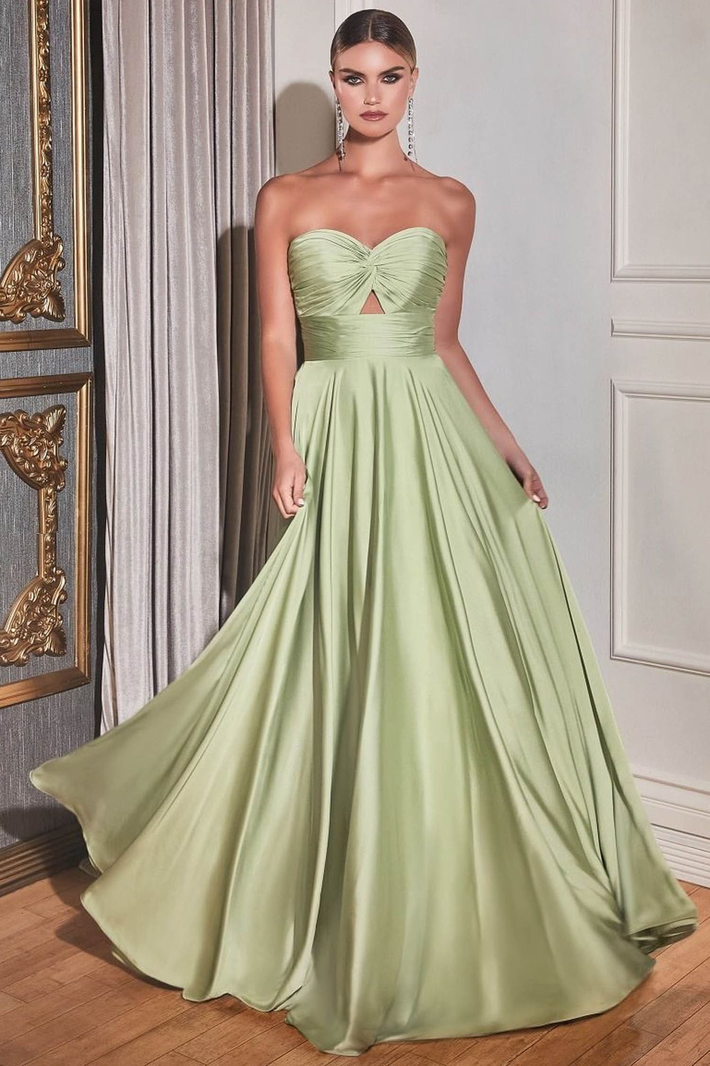 A woman models a strapless light sage green ball gown with a sweetheart neckline, a twisted bodice detail, and a full flowing skirt in an elegant indoor setting.