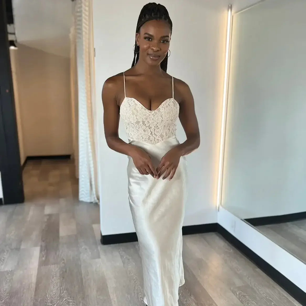 A woman stands elegantly, wearing a fitted lace and satin gown. She smiles while posing in a well-lit dressing room, featuring mirrors and wooden flooring.