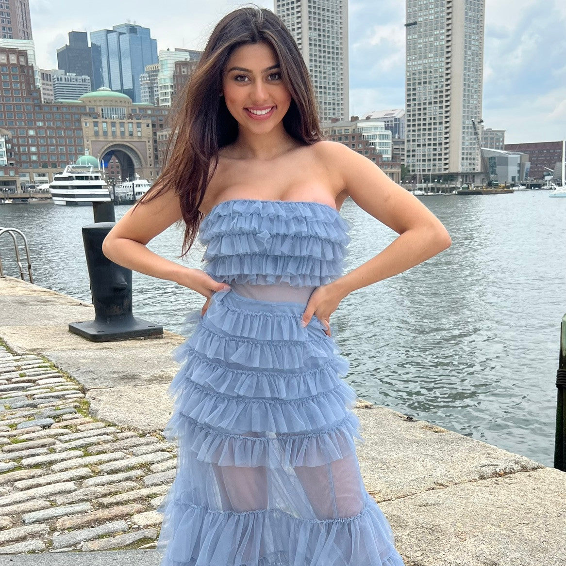 A woman stands on a cobblestone waterfront, smiling and posing in a blue ruffled dress. Behind her are modern buildings and boats on the water, under a cloudy sky.