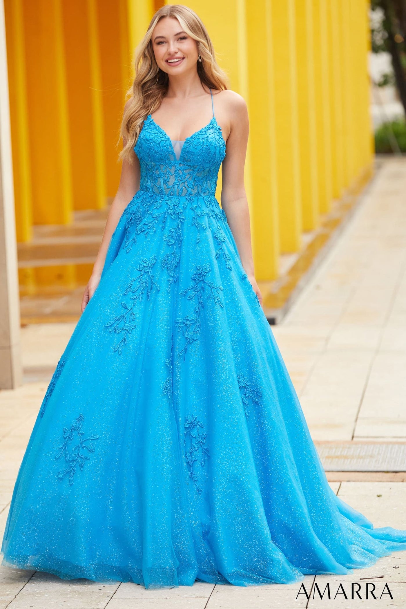 Woman wearing a blue evening gown with floral lace details against a yellow wall.