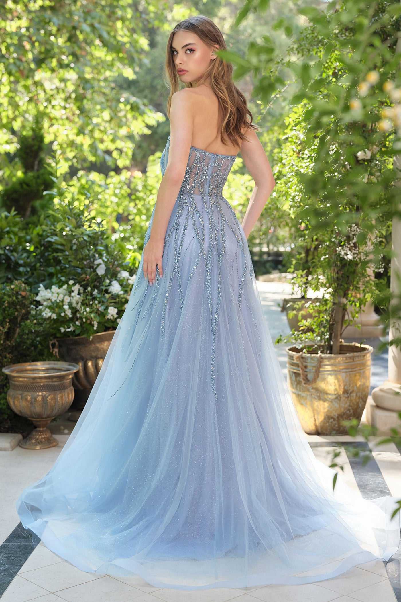 Woman in a light blue evening gown standing outdoors with greenery around.