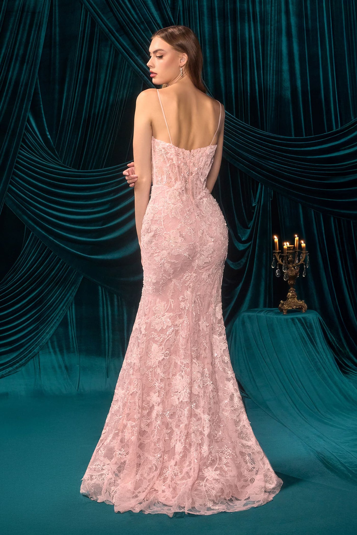 Woman in a pink lace evening gown standing against a dark teal curtain backdrop.