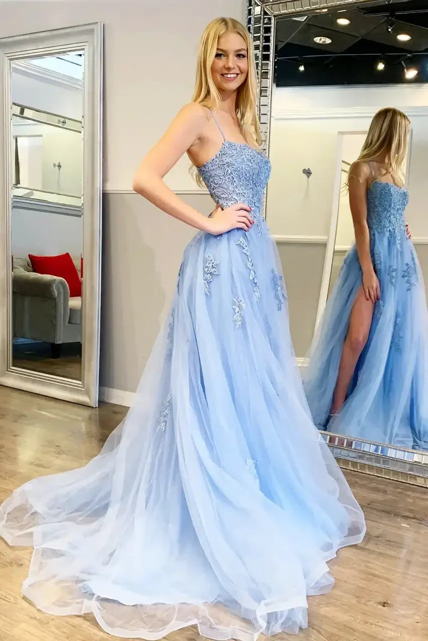 A woman poses in a flowing, light blue gown with floral embellishments, showcasing intricate details. She stands in a bright dressing room near a mirror, exuding confidence.