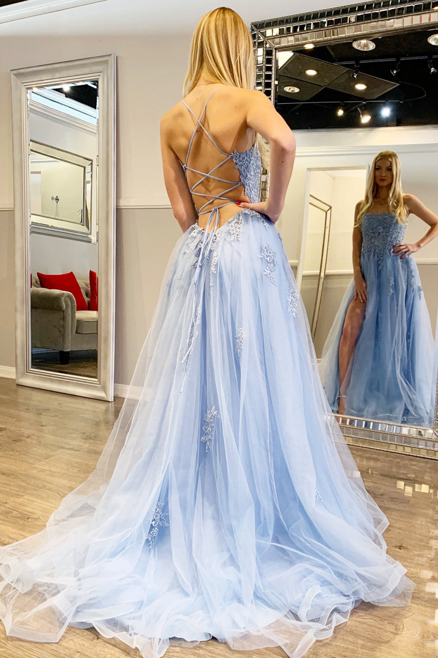 A woman models a flowing, light blue gown with intricate detailing. She poses with her back to the viewer, showcasing the dress's lace-up design in a bright, stylish boutique.