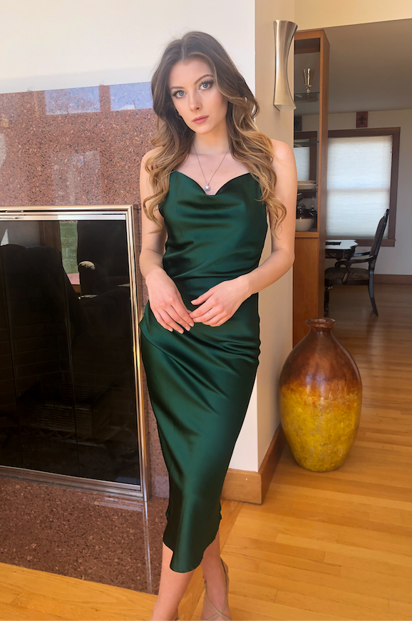 A woman stands elegantly in a form-fitting green satin dress, with one hand on her hip and the other relaxed. The background features a modern interior with wooden floors and a fireplace.