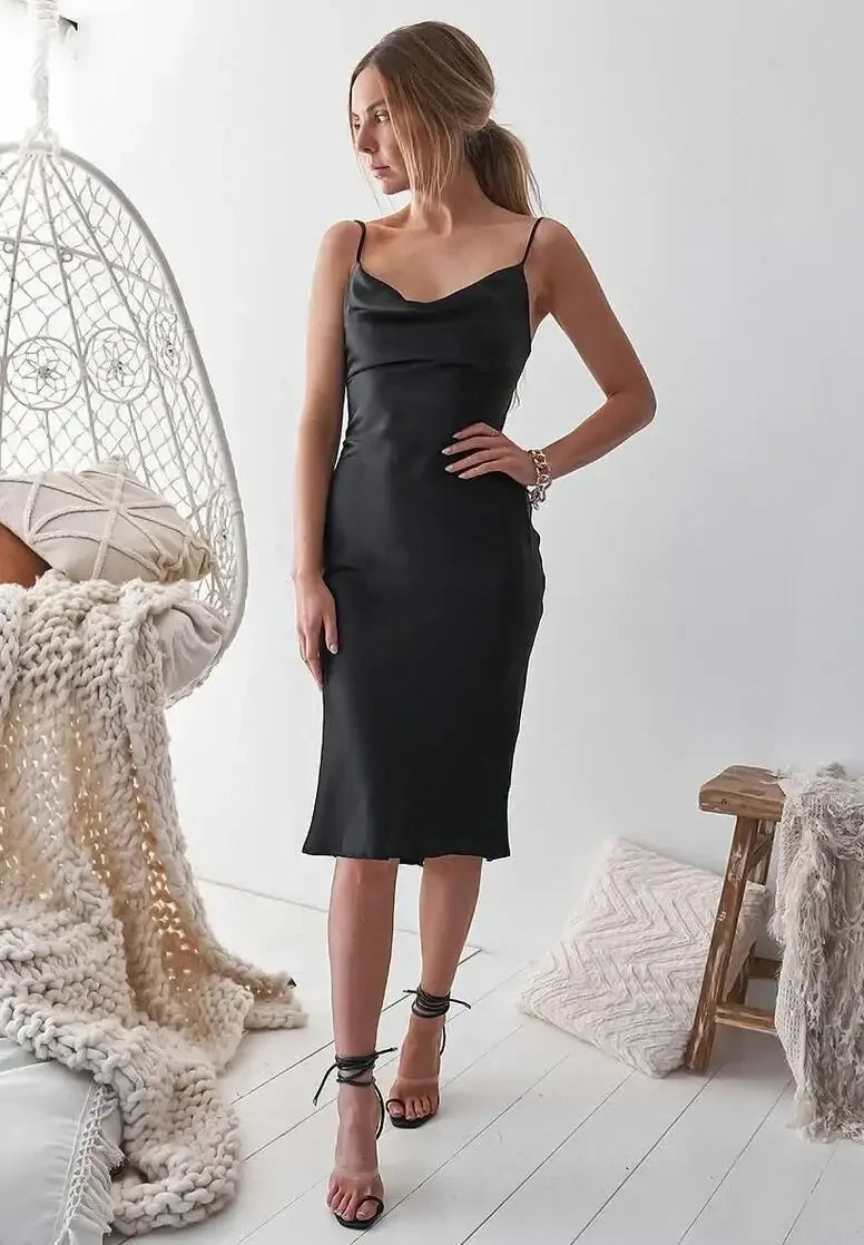 A woman in a sleek black satin dress stands elegantly, looking to the side. She is positioned in a bright, minimalistic space with a hanging chair and soft textiles nearby.