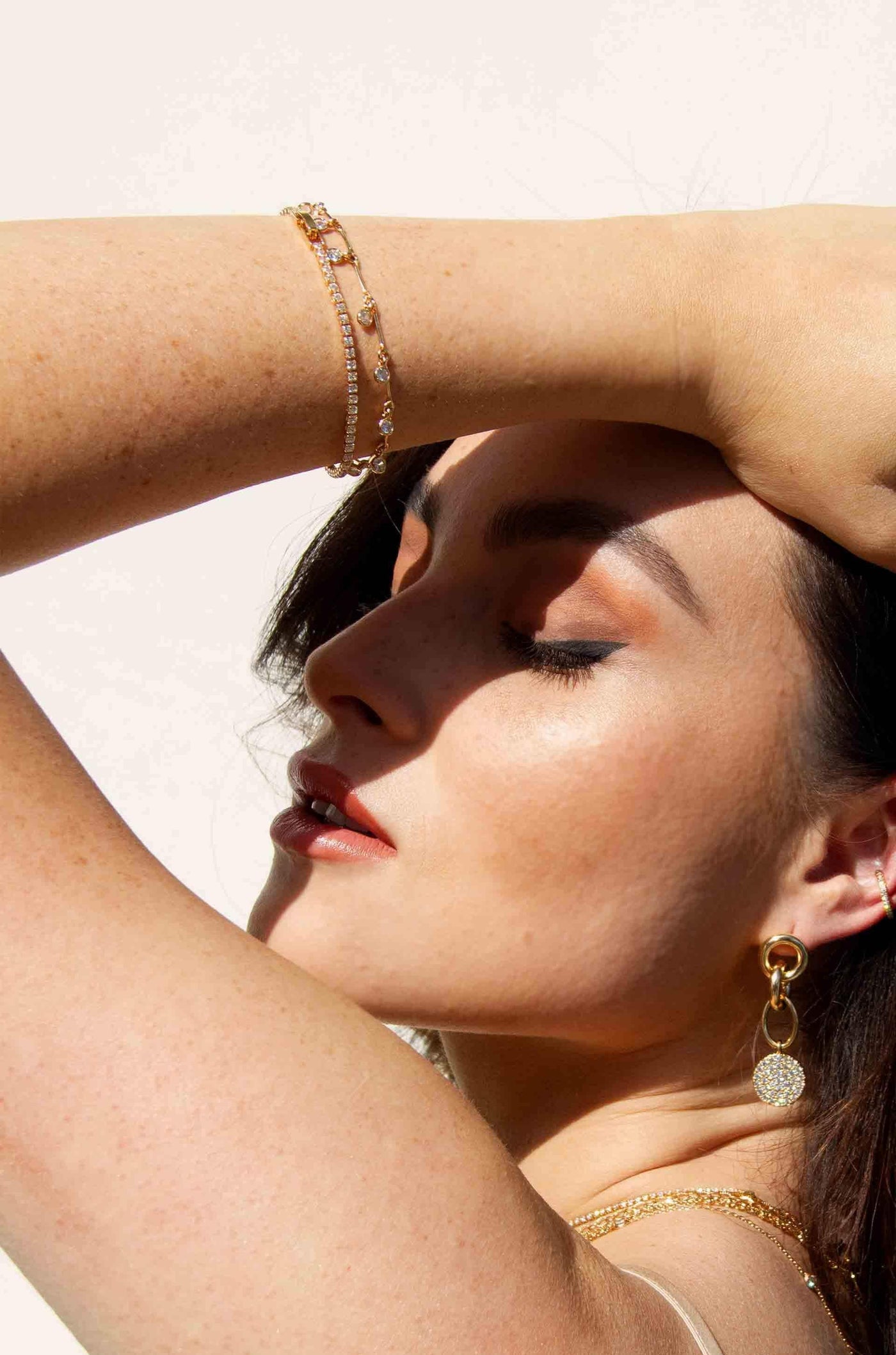 A woman with dark hair gracefully poses with her arm raised, showcasing delicate bracelets. Sunlight highlights her makeup and jewelry against a soft, light background.