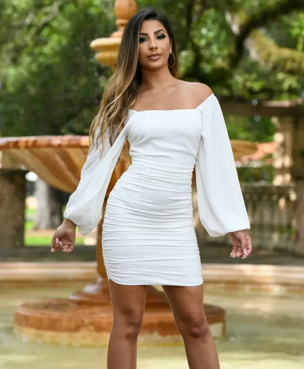 A woman in a fitted white dress with off-the-shoulder sleeves stands confidently in front of a decorative fountain surrounded by greenery, exuding elegance in a serene outdoor setting.