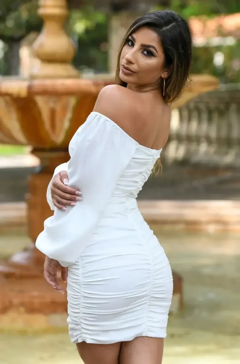 A woman in a fitted white off-the-shoulder dress poses gracefully by a fountain. She turns to look over her shoulder, surrounded by greenery and a blurred background of a serene park setting.