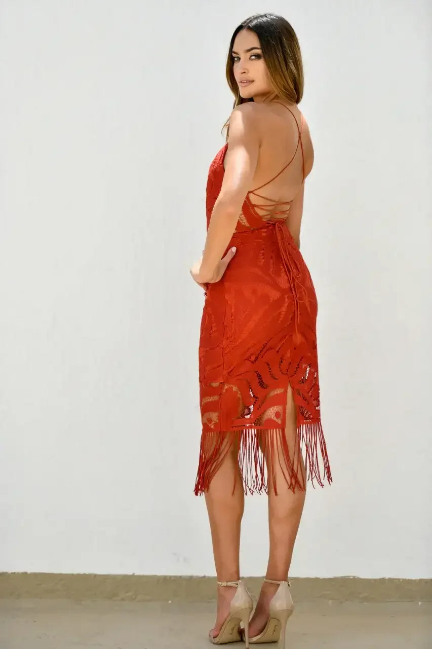 A woman poses in a red fringed dress, turning slightly to look back over her shoulder. She stands against a plain white wall, highlighting her elegant appearance.
