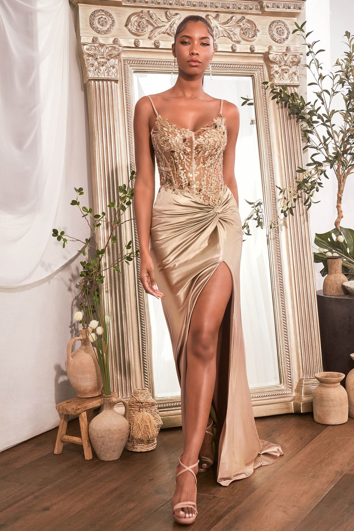 Woman in a gold evening gown standing in a decorated room with plants and mirrors.