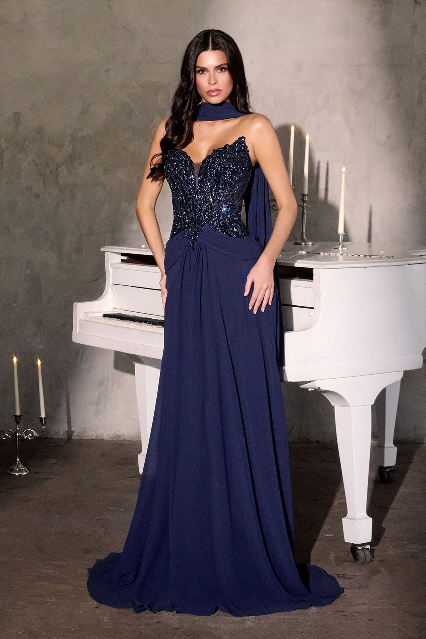 A woman models a floor-length navy blue evening gown with a beaded sequined bodice and a draped choker-style neckline, standing in front of a white grand piano against a textured gray wall.