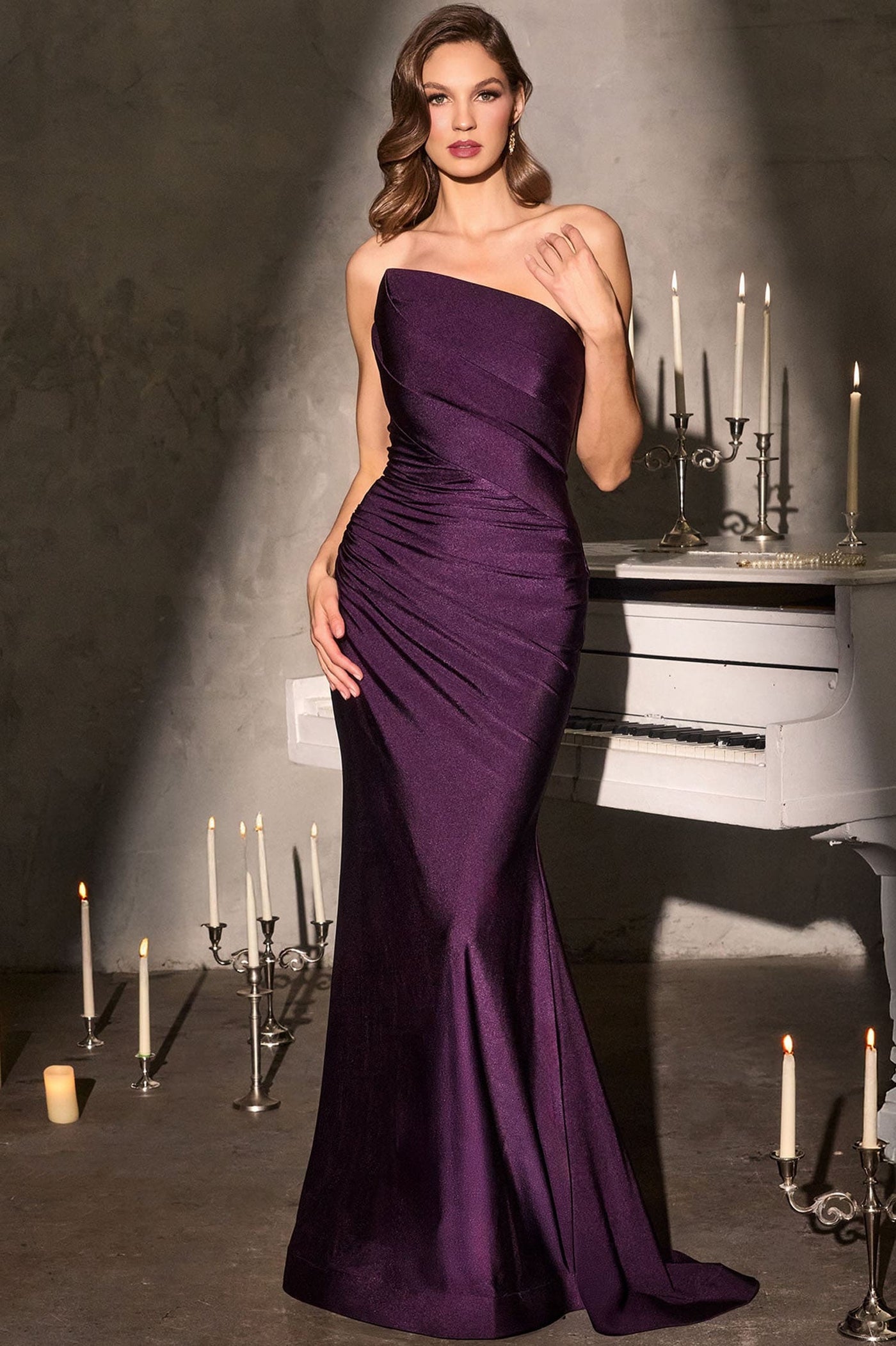 A woman models a strapless, floor-length mermaid gown in deep purple satin. The dress features a ruched bodice and a fitted skirt with a slight train.