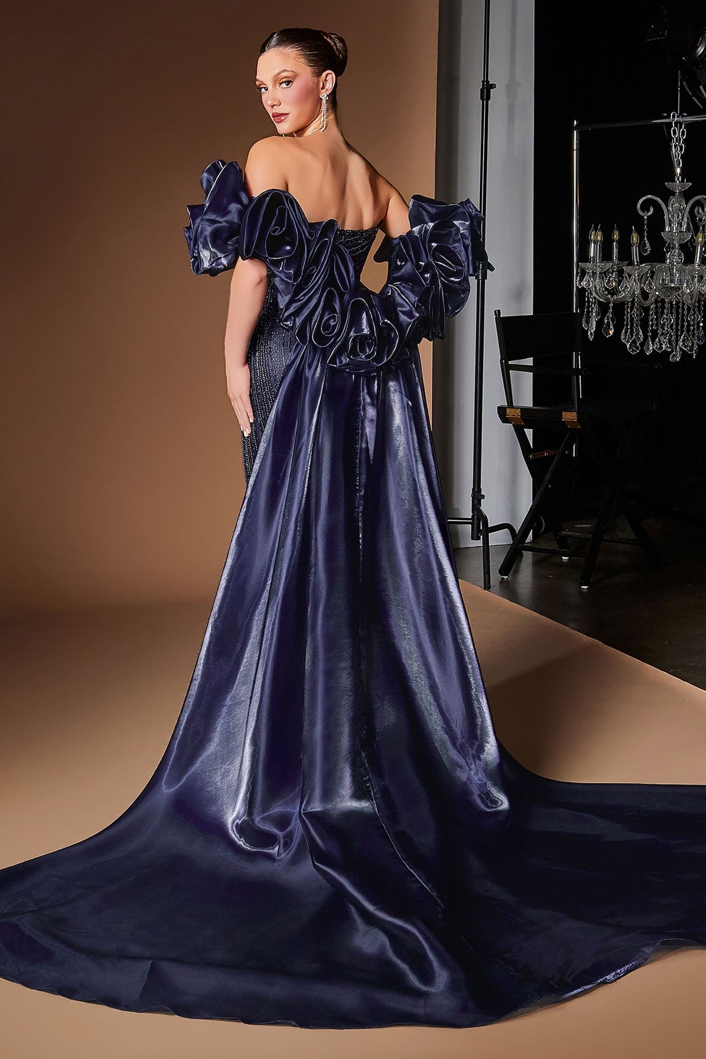 A woman modeling an off-the-shoulder navy blue gown with dramatic ruffled sleeves and a long satin train in a studio setting with a warm tan backdrop.