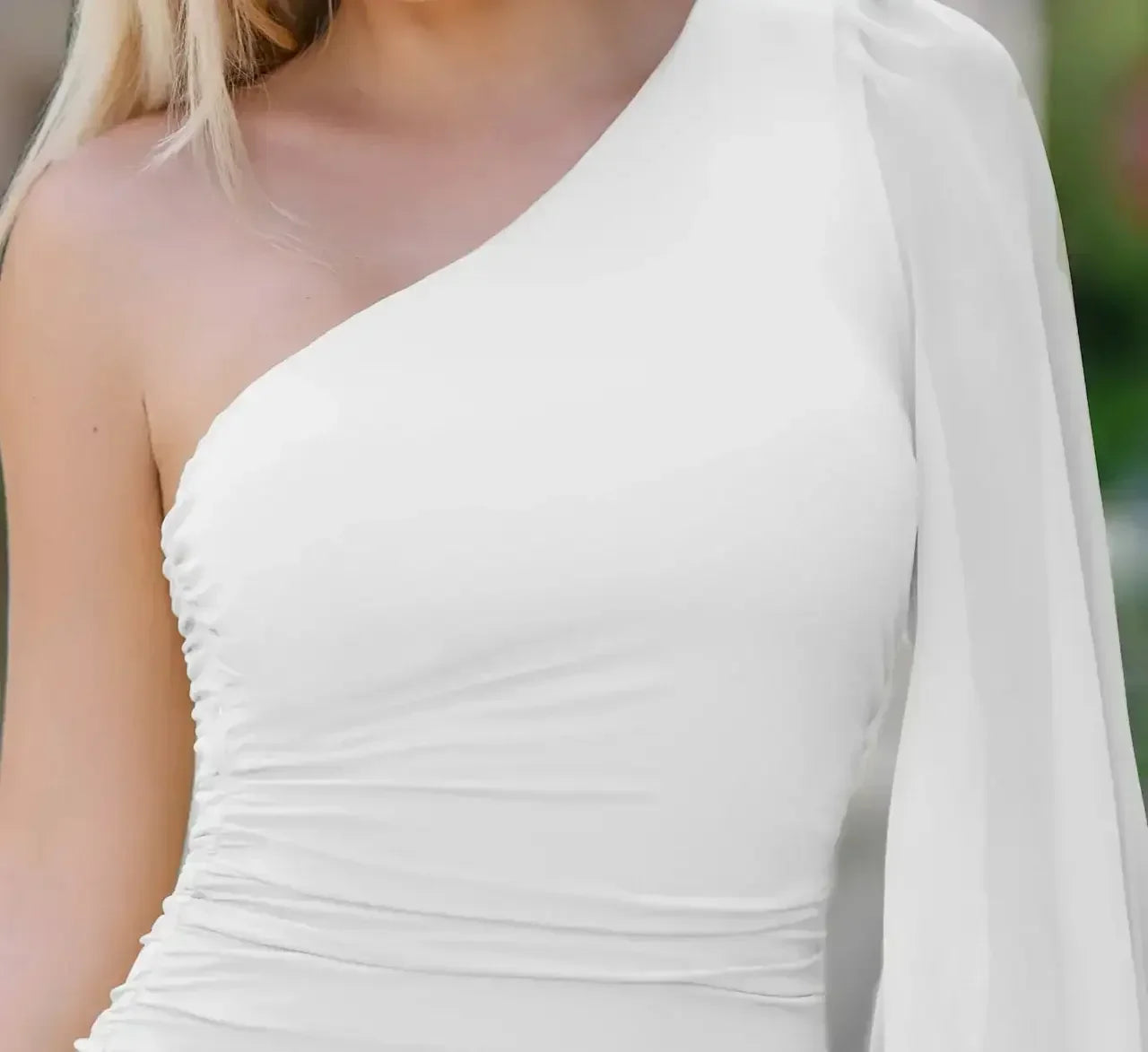 A person is wearing a stylish one-shoulder white outfit, showcasing a fitted bodice with gathered fabric. The background is softly blurred, suggesting an outdoor setting.