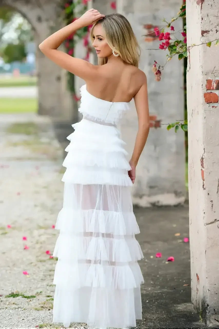 A woman stands gracefully, wearing a layered white dress with frills. She looks back, showcasing her hairstyle and earrings amidst a picturesque outdoor setting, with scattered flower petals on the ground.