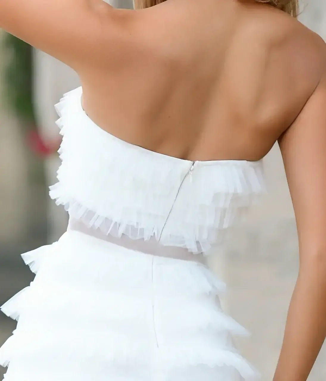 A white dress with layered, ruffled fabric features a strapless design and a visible zipper at the back, set against a softly blurred outdoor background.