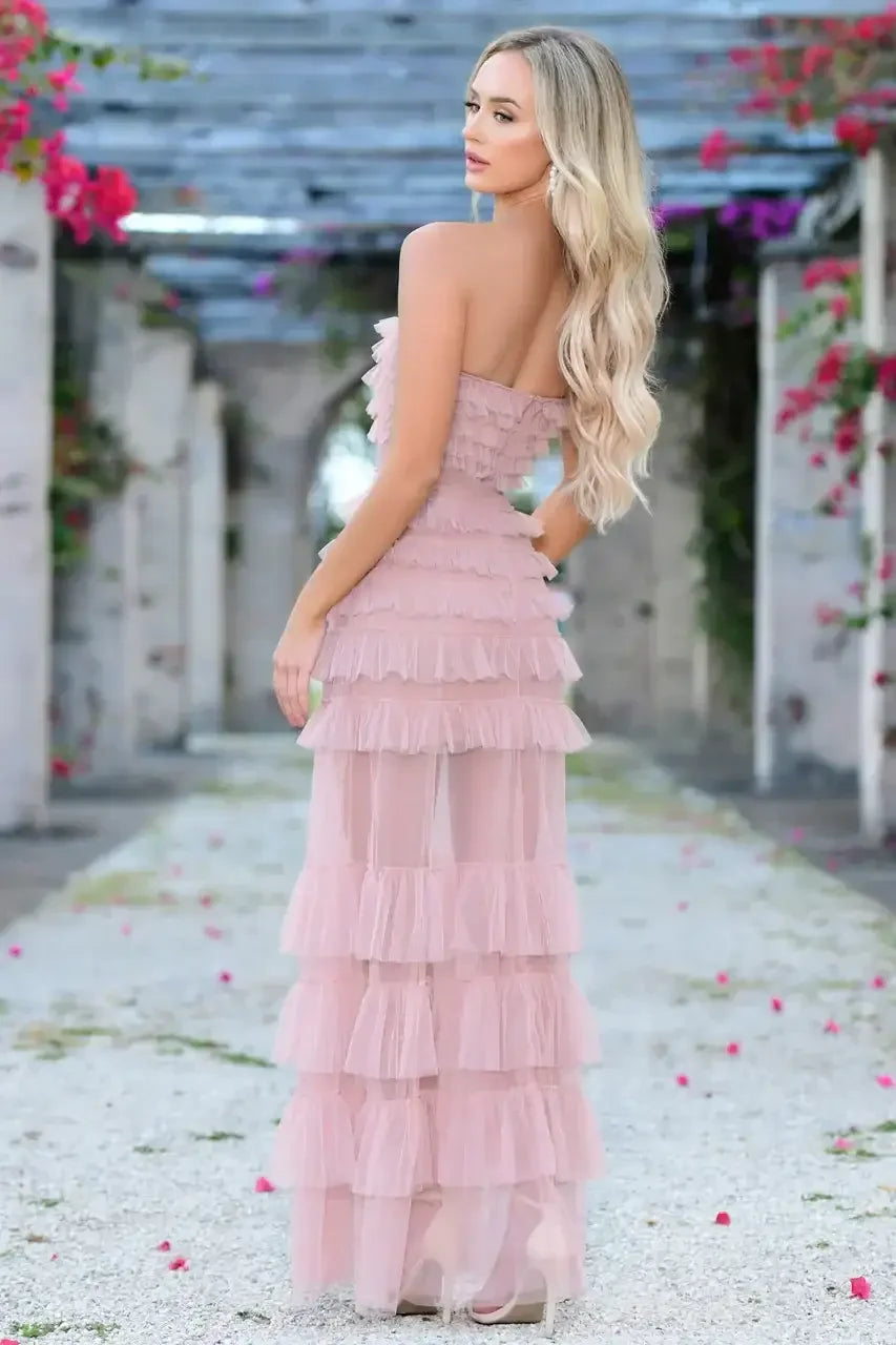 A woman is posed in a flowing, layered pink dress with ruffles, standing gracefully on a stone pathway adorned with scattered petals in a lush garden setting.