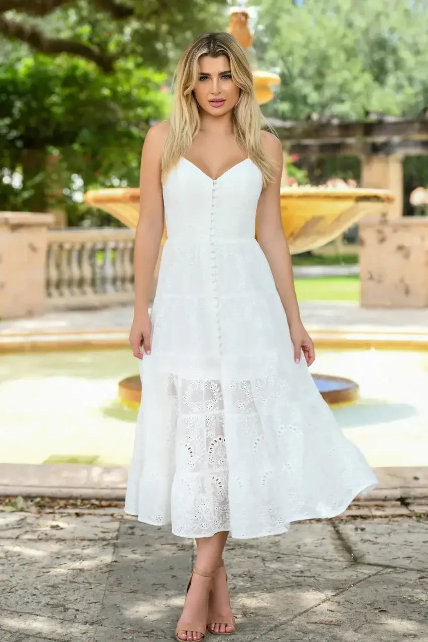 A woman stands elegantly in a flowing, white dress with lace details, holding her hands at her sides. The background features lush greenery and a stone fountain, enhancing a serene outdoor setting.