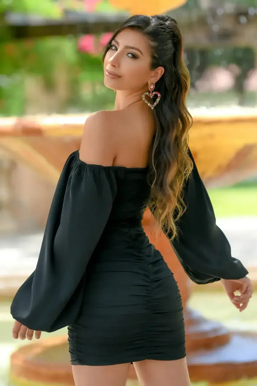 A woman in a black off-shoulder dress poses gracefully, smiling over her shoulder. She wears heart-shaped earrings, with a lush, floral garden backdrop and soft sunlight enhancing the scene.