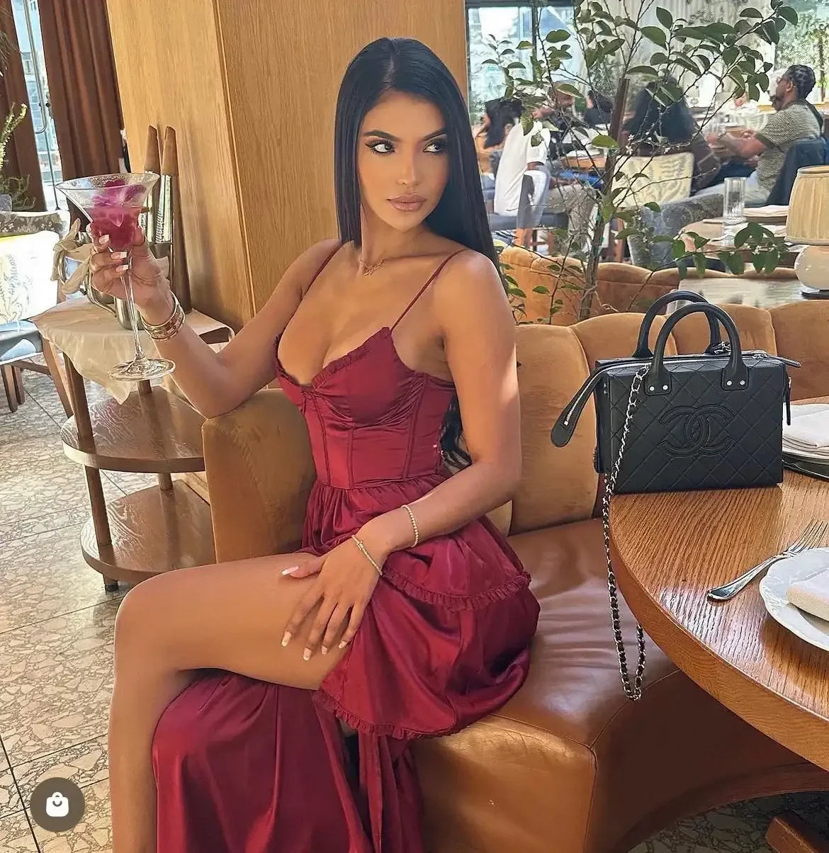 A woman in a red dress sits confidently at a table, holding a cocktail. Nearby, a black designer handbag rests on the table, surrounded by greenery and patrons in a stylish dining environment.