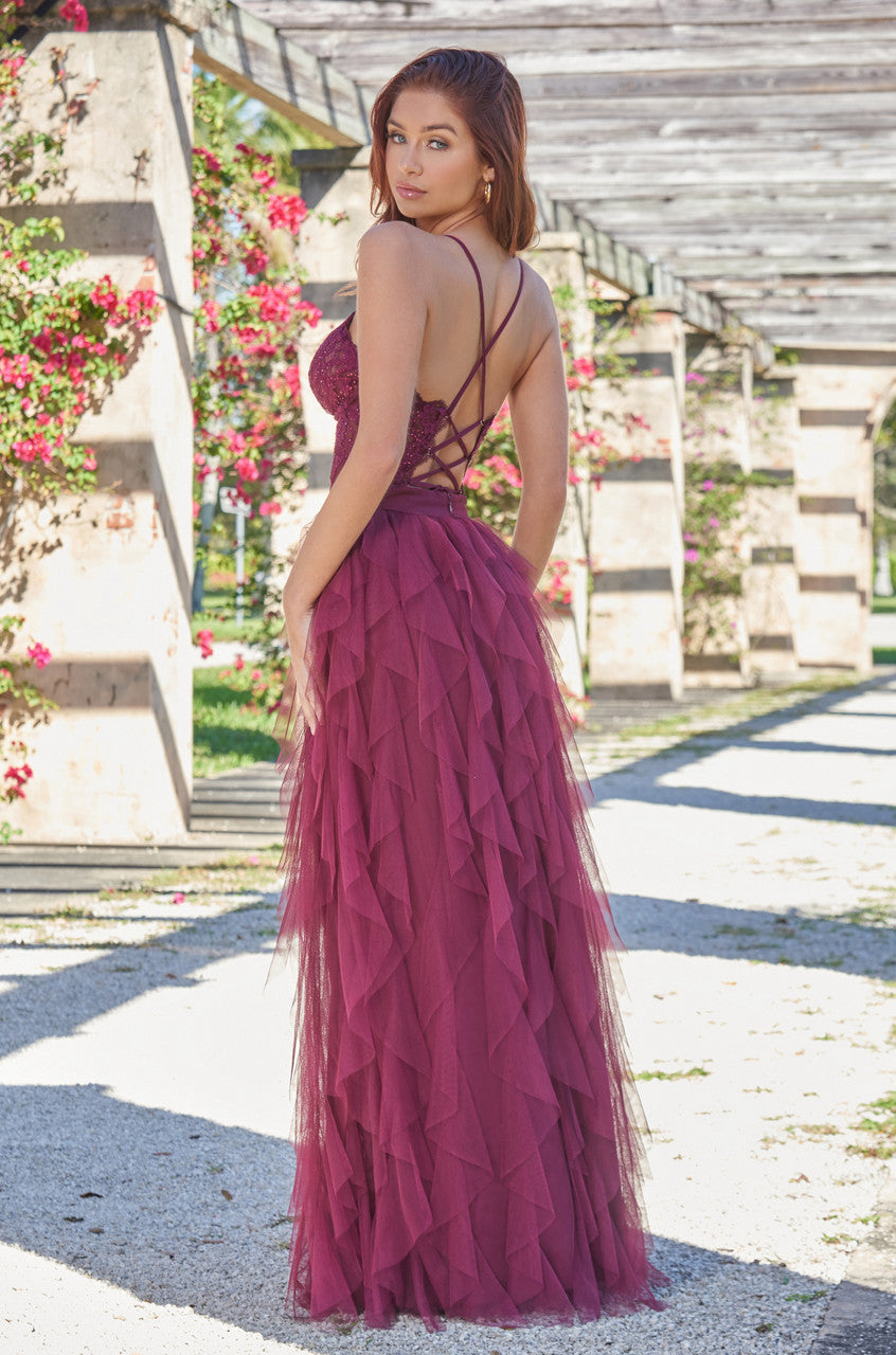 A woman in a burgundy, tiered gown stands gracefully in a garden. She poses with one hand on her hip, showcasing the dress's intricate back design amidst blooming flowers and stone archways.