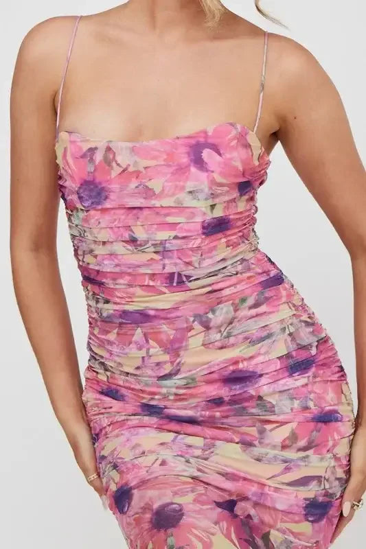 A woman is wearing a fitted, floral-patterned dress with ruching, predominantly in shades of pink and purple, against a light background, showcasing a stylish and elegant look.