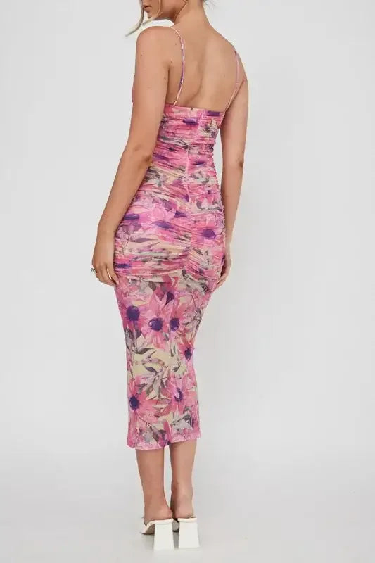 A person stands wearing a floral-printed, ruched dress with thin straps. The dress features pink hues and tropical patterns, in a minimalistic studio setting with a plain white background.