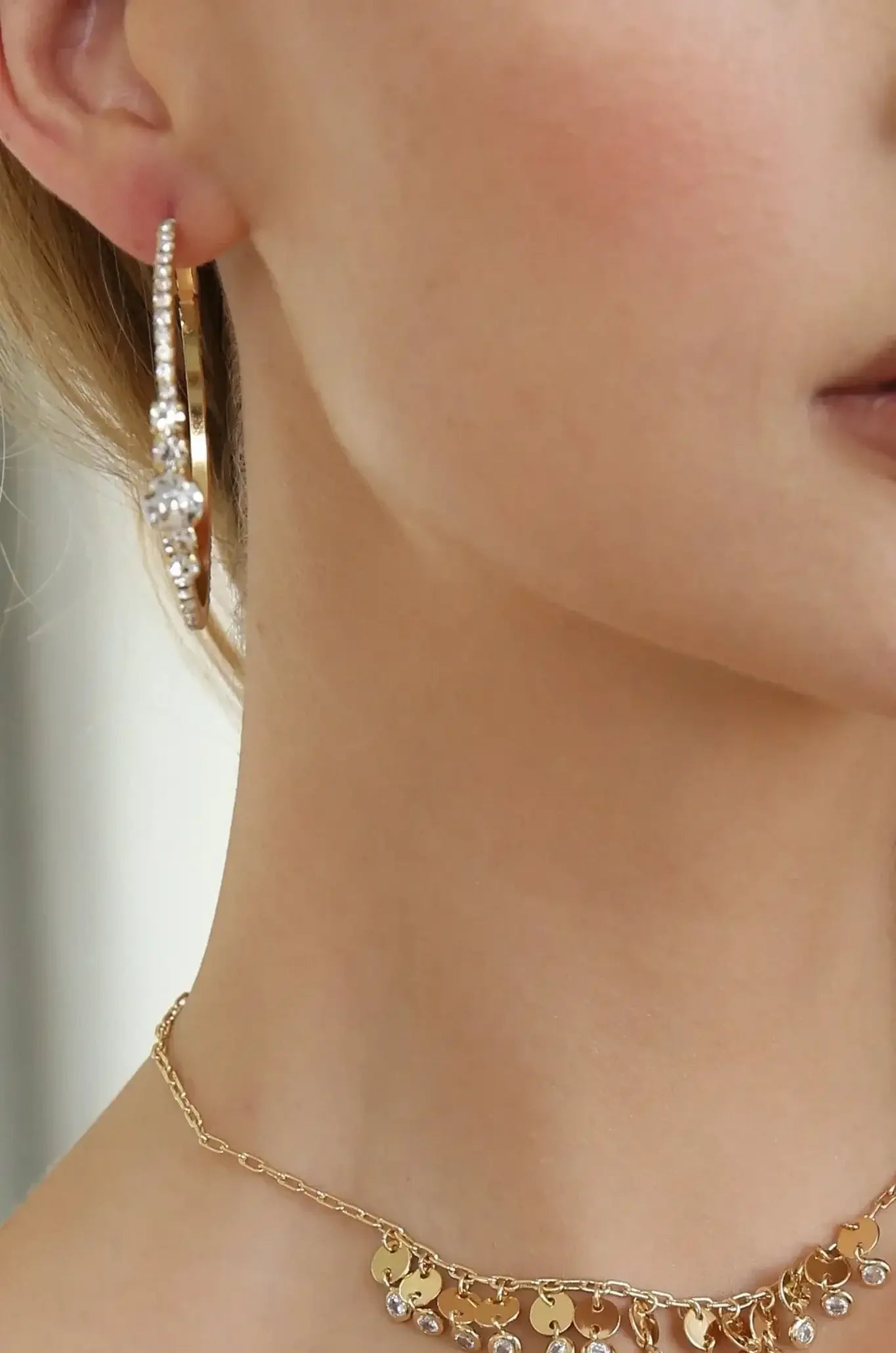 A close-up of a woman’s neck features a gold necklace with a pendant and a large, ornate gold hoop earring adorned with sparkling stones, set against a light background.